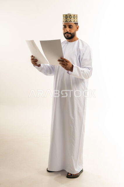 القراءة والمطالعة ودراسة المشروع، بورتريه لرجل عربي خليجي عماني يرتدي الدشداشة والكمة يمسك بيده مجموعة أوراق يقوم بتصفحها بإيماءات التركيز والتمعن، قضاء وقت الفراغ في التثقيف والتعليم، مفهوم التعلم الذاتي، الجسم بالطول الكامل، خلفية بيضاء