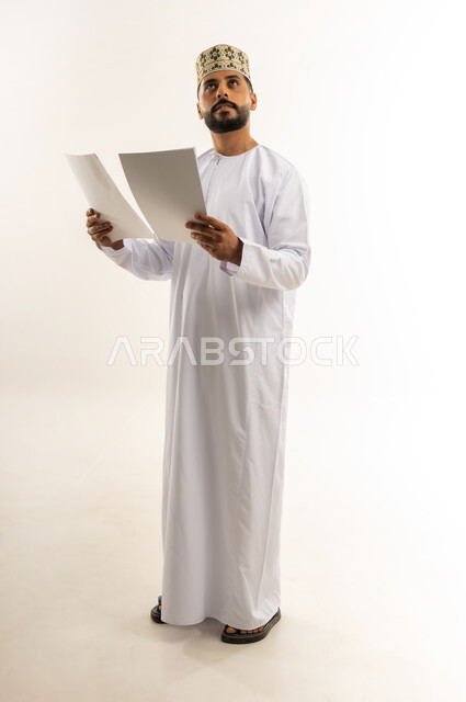 محاولة تذكر أمر ما، مفهوم التعلم الذاتي، القراءة والمطالعة ودراسة المشروع، بورتريه لرجل عربي خليجي عماني يرتدي الدشداشة والكمة يمسك بيده مجموعة أوراق ينظر إلى الأعلى بإيماءات التفكير، قضاء وقت الفراغ في التثقيف والتعليم، الجسم بالطول الكامل، خلفية بيضاء