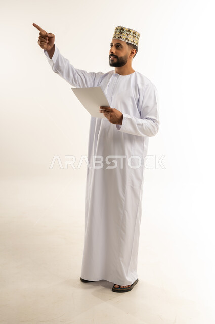 الإشارة بإصبع السبابة إلى شيء ما في الأعلى، مفهوم التعلم الذاتي، القراءة والمطالعة ودراسة المشروع، بورتريه لرجل عربي خليجي عماني يرتدي الدشداشة والكمة يمسك بيده مجموعة أوراق، قضاء وقت الفراغ في التثقيف والتعليم، الجسم بالطول الكامل، خلفية بيضاء