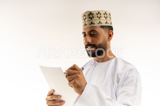 Daily task writing tools, using white paper to record important notes and information, close-up portrait of an Arab Gulf Omani man wearing a dishdasha and a turban holding a paper and pen in his hand writing something with gestures of concentration and contemplation, white background