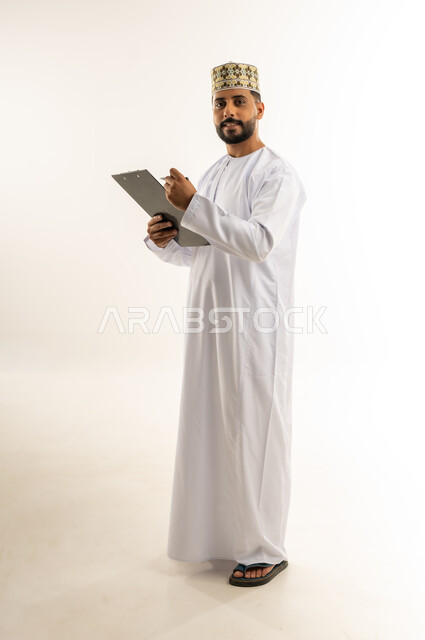 أدوات تدوين المهام اليومية، تسجيل الملاحظات والمعلومات المهمة, بورتريه لرجل عربي خليجي عماني يرتدي الدشداشة والكمة يمسك بيديه سنادة ورق سوداء وقلم يقوم بكتابة شيء ما بنظر إلى الكاميرا بإيماءات السرور، الجسم بالطول الكامل، خلفية بيضاء
