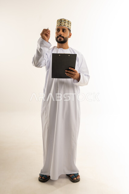 تسجيل الملاحظات والمعلومات المهمة, بورتريه لرجل عربي خليجي عماني يرتدي الدشداشة والكمة يمسك بيديه سنادة ورق سوداء وقلم ينظر إليه بإيماءات التركيز والتمعن، أدوات تدوين المهام اليومية، الجسم بالطول الكامل، خلفية بيضاء
