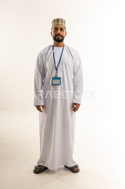 الوقوف باستقامة والنظر إلى الكاميرا، بورتريه لرجل عربي خليجي عماني يرتدي الدشداشة والكمة يضع الهوية الشخصية حول رقبته ينظر بإيماءات الجدية والثقة بالنفس, مفهوم الرجولة والأناقة، الاهتمام بالمظهر الخارجي, الجسم بالطول الكامل, خلفية بيضاء