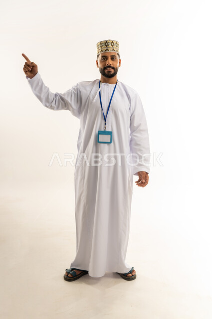 الإشارة لموضع معين, بورتريه لرجل عربي خليجي عماني يرتدي الدشداشة والكمة يضع الهوية الشخصية حول رقبته يشير بإصبع السبابة إلى شيء ما في الأعلى ينظر إلى الكاميرا بإيماءات الثقة بالنفس, الاهتمام بالمظهر الخارجي, الجسم بالطول الكامل, خلفية بيضاء