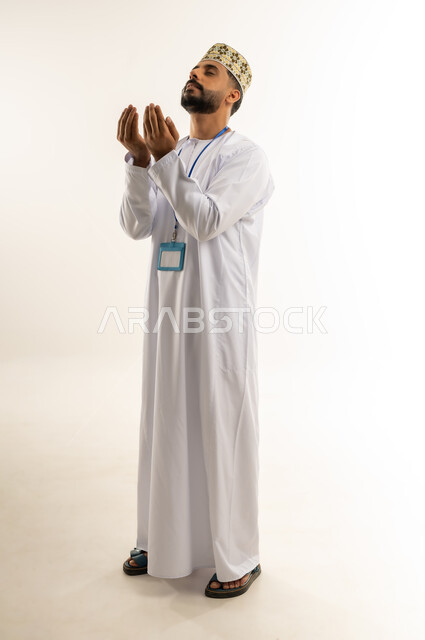 The concept of worship and drawing closer to God Almighty, striving in supplication, obedience and remembrance, portrait of an Arab Gulf Omani man wearing a dishdasha and a kummah, placing his personal ID around his neck, raising his hands up with expressions of supplication and reverence to the Lord of the Worlds, full-length body, white background