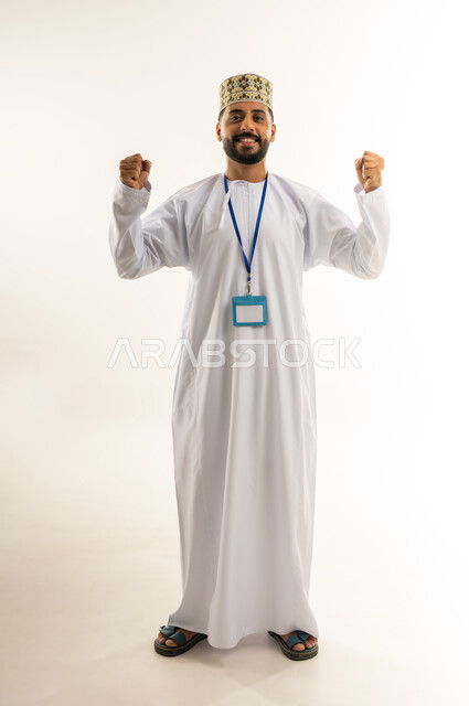 رفع قبضتي اليد للأعلى بإيماءات الفوز والانتصار، بورتريه لرجل عربي خليجي عماني مبتسم يرتدي الدشداشة والكمة يضع الهوية الشخصية حول رقبته ينظر إلى الكاميرا بتعابير السرور والفخر، إنجاز المهمة المطلوبة، الجسم بالطول الكامل, خلفية بيضاء