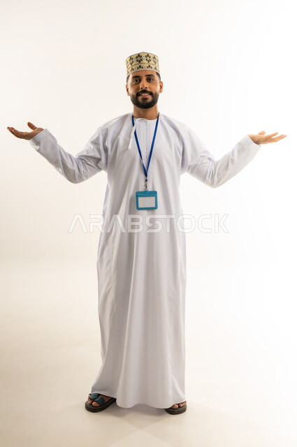 التردد وعدم الثقة بخصوص أمر معين، بورتريه لرجل عربي خليجي عماني يرتدي الدشداشة والكمة يضع الهوية الشخصية على رقبته ينظر إلى الكاميرا يرفع يديه يفتح كفيه بإيماءات عدم التأكد, الشعور بالقلق وعدم المعرفة، الحيرة باتخاذ القرار الصحيح، الجسم بالطول الكامل، خلفية بيضاء