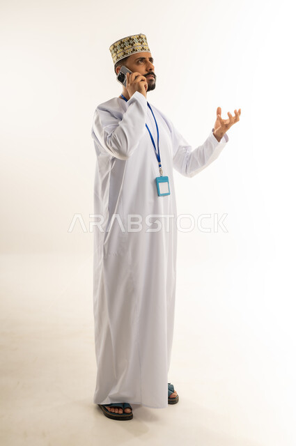 سماع الأخبار غير الجيدة، استخدام جهاز تقني حديث، التواصل مع الأهل والأصدقاء، بورتريه لرجل عربي خليجي عماني يرتدي الدشداشة والكمة يضع الهوية الشخصية على رقبته يقوم بإجراء مكالمة هاتفية عبر الهاتف المحمول يرفع يده بإيماءات الغضب، الجسم بالطول الكامل، خلفية بيضاء