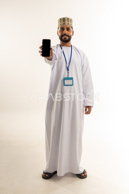 عرض تطبيقات الجوال، شاشة سوداء فارغة، استخدام جهاز تقني حديث، بورتريه لرجل عربي خليجي عماني يرتدي الدشداشة والكمة يضع الهوية الشخصية على رقبته يمسك بيده الهاتف المحمول ينظر إلى الكاميرا بإيماءات السرور، الجسم بالطول الكامل، خلفية بيضاء