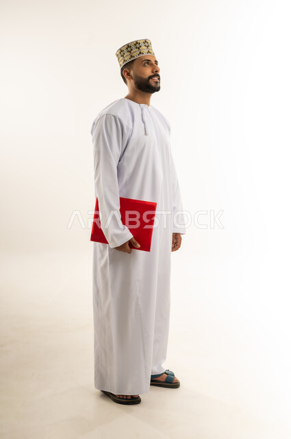 دراسة جدوى العمل، مراجعة نتيجة الاجتماع، بورتريه لرجل عربي خليجي عماني يرتدي الدشداشة والكمة يحمل بيده ملف باللون الأحمر ينظر إلى شيء ما في الأعلى بإيماءات الجدية والثقة، إعداد التقرير اليومي، الجسم بالطول الكامل، خلفيه بيضاء