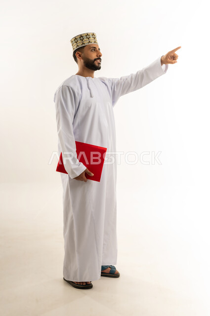الإشارة بإصبع السبابة إلى شيء ما في الأعلى، إعداد التقرير اليومي، دراسة جدوى العمل، مراجعة نتيجة الاجتماع، بورتريه لرجل عربي خليجي عماني يرتدي الدشداشة والكمة يحمل بيده ملف باللون الأحمر ينظر إلى مكان معين بإيماءات الجدية والثقة، الجسم بالطول الكامل، خلفيه بيضاء
