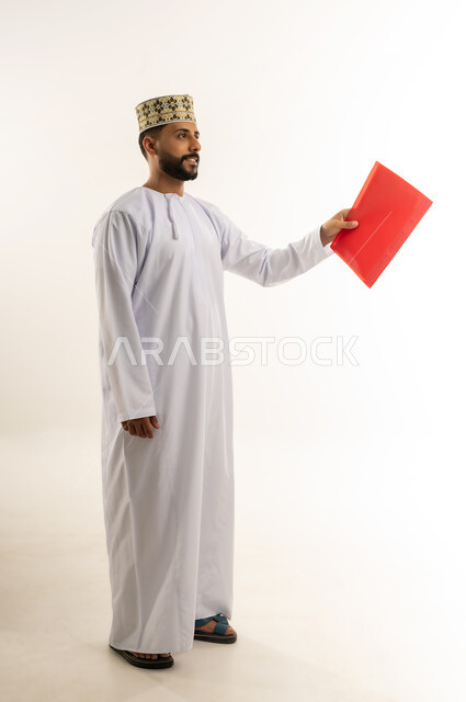 دراسة جدوى العمل، مراجعة نتيجة الاجتماع، بورتريه لرجل عربي خليجي عماني يرتدي الدشداشة والكمة يحمل بيده ملف باللون الأحمر يقوم بتقديمه إلى شخص ما ينظر إلى مكان معين بإيماءات الجدية والثقة، تدوين الملاحظات وإعداد التقرير اليومي، الجسم بالطول الكامل، خلفيه بيضاء
