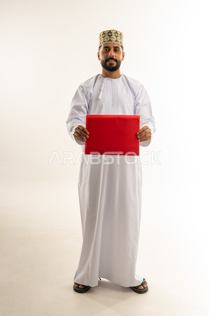 تدوين الملاحظات وإعداد التقرير اليومي، مراجعة نتيجة الاجتماع، بورتريه لرجل عربي خليجي عماني يرتدي الدشداشة والكمة يحمل بيديه ملف باللون الأحمر ينظر إلى الكاميرا بايماءات الجدية والثقة، دراسة جدوى العمل، الجسم بالطول الكامل، خلفيه بيضاء