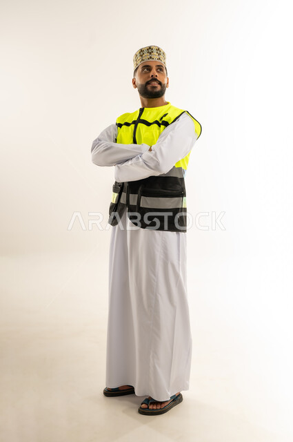 Career and job for youth in the Sultanate of Oman, concept of architectural constructions, portrait of an Arab Gulf Omani engineer wearing a dishdasha, a turban and a protective vest standing with crossed hands looking at something above with gestures of self-confidence, supervising engineering projects, full-length body, white background