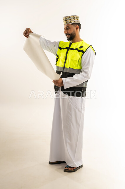 Concept of engineering and architectural construction, portrait of an Arab Gulf Omani engineer wearing a dishdasha, a cloak and a protective vest holding a blueprint and looking at it with gestures of concentration and contemplation, profession and job for young people in the Sultanate of Oman, working in the engineering sector, full body, white background