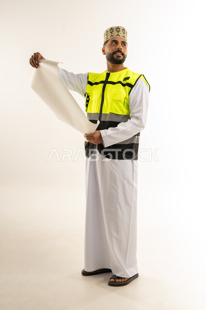 Career and job for young people in the Sultanate of Oman, supervising engineering project plans, concept of architectural constructions, portrait of an Arab Gulf Omani engineer wearing a dishdasha, a turban and a protective vest holding a planner in his hands looking at something, working in the field of engineering, full body, white background