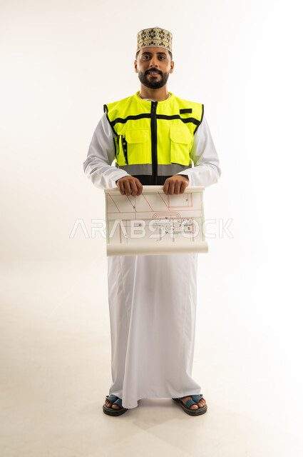 الإشراف على مخططات المشاريع الهندسية، مفهوم الانشاءات المعمارية، بورتريه لمهندس عربي خليجي عماني يرتدي الدشداشة والكمة وسترة الحماية يمسك بيديه المخطط ينظر إلى الكاميرا بإيماءات الثقة بالنفس، مهنة ووظيفة للشباب في سلطنة عمان، الجسم بالطول الكامل، خلفية بيضاء