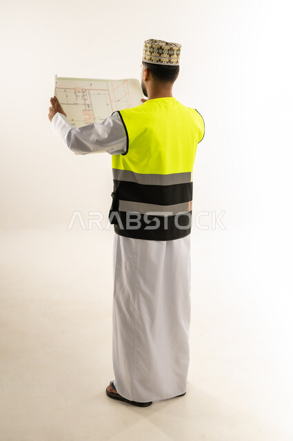 مفهوم الانشاءات المعمارية، بورتريه من الخلف لمهندس عربي خليجي عماني يرتدي الدشداشة والكمة وسترة الحماية يمسك بيديه المخطط، العمل في مجال الهندسة، مهنة ووظيفة للشباب في سلطنة عمان، الإشراف على مخططات المشاريع الهندسية، الجسم بالطول الكامل، خلفية بيضاء