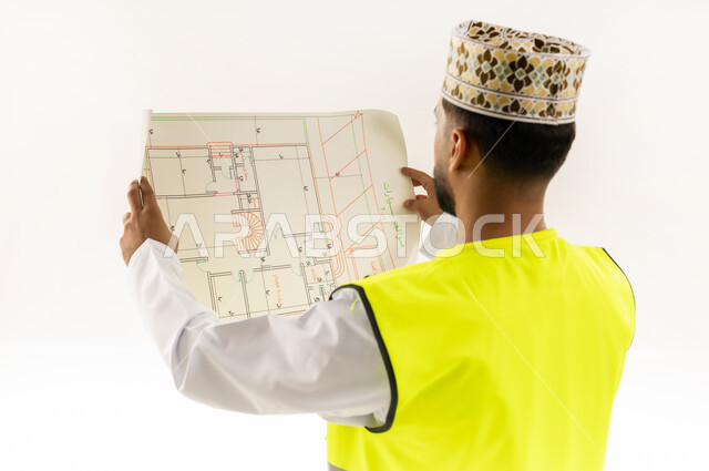 الإشراف على مخططات المشاريع الهندسية، مفهوم الانشاءات المعمارية، بورتريه مقرب من الخلف لمهندس عربي خليجي عماني يرتدي الدشداشة والكمة وسترة الحماية يمسك بيديه المخطط، العمل في مجال الهندسة، مهنة ووظيفة للشباب في سلطنة عمان، خلفية بيضاء