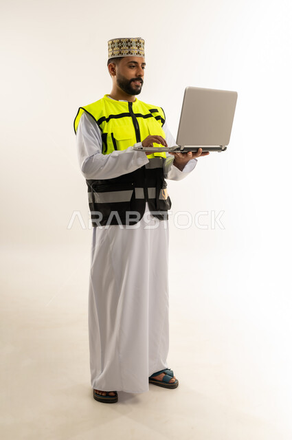 الإشراف على العمل باستخدام التكنولوجيا المتطورة، الاعتماد على التقنية الحديثة بالقطاع الهندسي، بورتريه لمهندس عربي خليجي عماني يرتدي الدشداشة والكمة وسترة الحماية يمسك الحاسوب المحمول يقوم بمتابعة أعماله عن بعد، الجسم بالطول الكامل، خلفية بيضاء