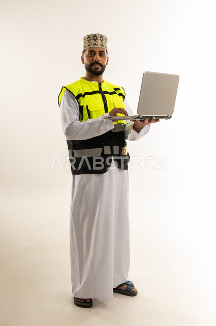 الاعتماد على التقنية الحديثة بالقطاع الهندسي، بورتريه لمهندس عربي خليجي عماني يرتدي الدشداشة والكمة وسترة الحماية يمسك الحاسوب المحمول ينظر إلى الكاميرا بإيماءات الثقة، الإشراف على العمل عن بعد باستخدام التكنولوجيا المتطورة، الجسم بالطول الكامل، خلفية بيضاء