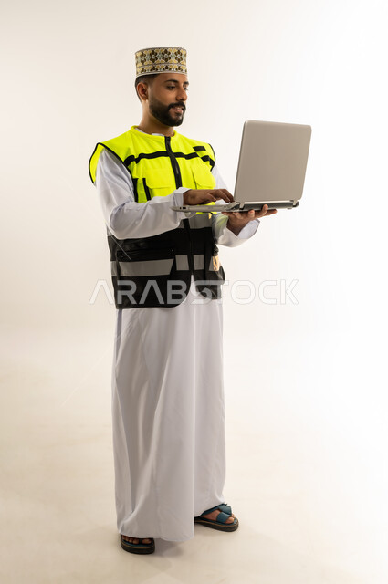 الإشراف على العمل باستخدام التكنولوجيا المتطورة، الاعتماد على التقنية الحديثة بالقطاع الهندسي، بورتريه لمهندس عربي خليجي عماني يرتدي الدشداشة والكمة وسترة الحماية يمسك الحاسوب المحمول يقوم بمتابعة أعماله عن بعد، الجسم بالطول الكامل، خلفية بيضاء
