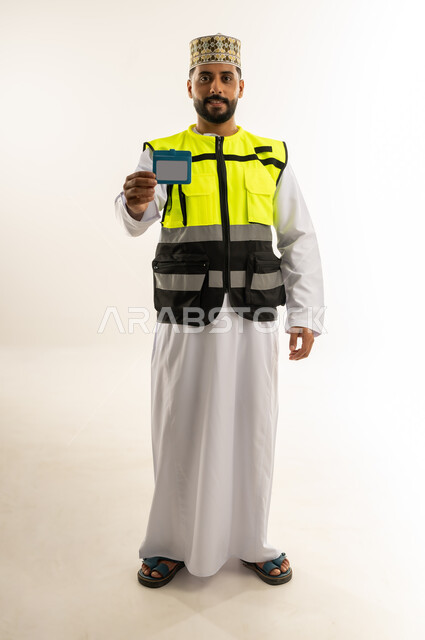 مفهوم الهندسة والانشاءات المعمارية، بورتريه لمهندس عربي خليجي عماني يرتدي الدشداشة والكمة وسترة الحماية يعرض هوية شخصية ينظر إلى الكاميرا بايماءات الثقة بالنفس، العمل في القطاع الهندسي، مهنة ووظيفة للشباب في سلطنة عمان، الجسم بالطول الكامل، خلفية بيضاء