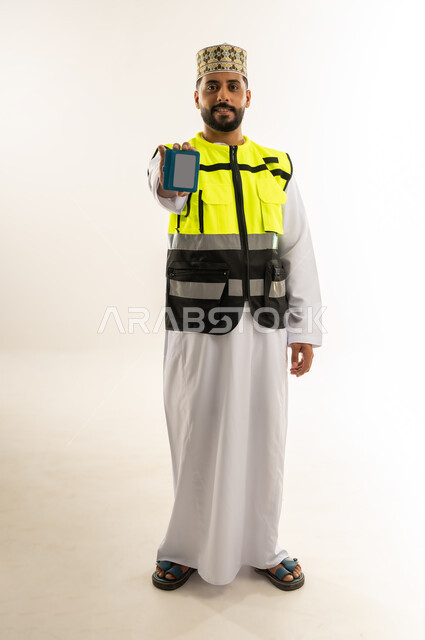 Working in the engineering sector, concept of engineering and architectural construction, portrait of an Arab Gulf Omani engineer wearing a dishdasha, a kimono and a protective vest, displaying a personal identity, looking at the camera with gestures of self-confidence, profession and job for young people in the Sultanate of Oman, full-length body, white background