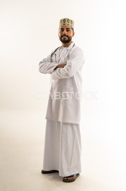 النظر إلى الكاميرا بإيماءات الثقة بالنفس، العمل في القطاع الطبي في سلطنة عمان، مفهوم الطب والرعاية الصحية، بورتريه لطبيب عربي خليجي عماني يرتدي الدشداشة والكمة والزي الخاص بالمهنة يقف بأيدي متقاطعة، الجسم بالطول الكامل، خلفية بيضاء