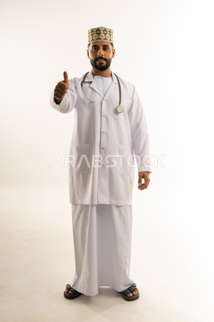 علامة الجودة والامتياز، العمل في القطاع الطبي في سلطنة عمان، مفهوم الطب والرعاية الصحية، بورتريه لطبيب عربي خليجي عماني يرتدي الدشداشة والكمة والزي الخاص بالمهنة ينظر إلى الكاميرا يرفع إصبع الإبهام يإيماءات الإعجاب والقبول، الجسم بالطول الكامل، خلفية بيضاء