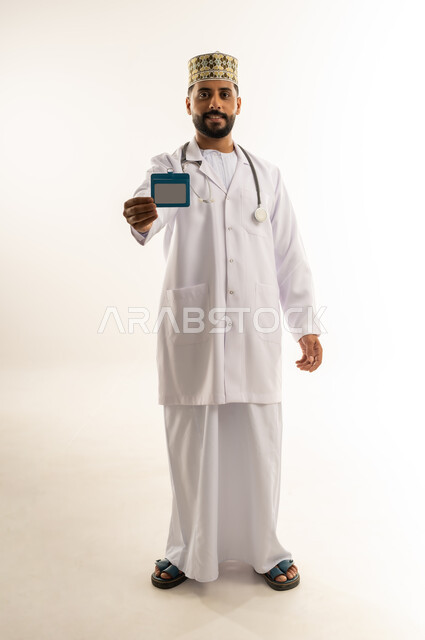العمل في القطاع الطبي في سلطنة عمان، مفهوم الطب والرعاية الصحية، بورتريه لطبيب عربي خليجي عماني يرتدي الدشداشة والكمة والزي الخاص بالمهنة يعرض هوية شخصية مستطيلة باللون الأبيض ينظر إلى الكاميرا يإيماءات الثقة بالنفس، الجسم بالطول الكامل، خلفية بيضاء