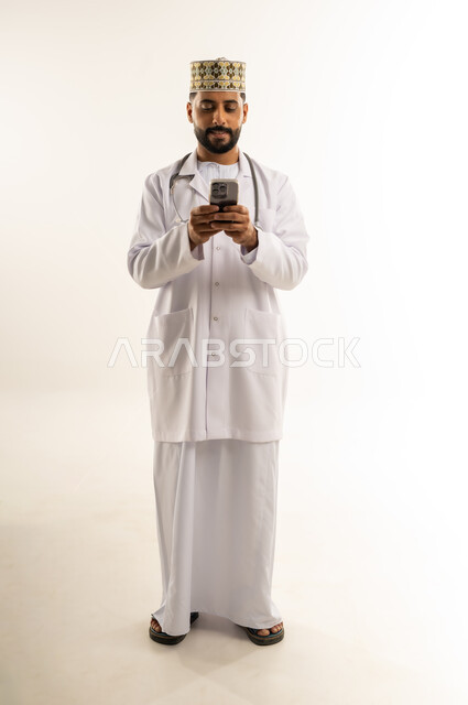 العمل في القطاع الطبي في سلطنة عمان، تقديم الاستشارة للمرضى عن بعد من خلال الجوال, بورتريه لطبيب عربي خليجي عماني يرتدي الدشداشة والكمة والزي الخاص بالمهنة يستخدم الهاتف المحمول، استخدام جهاز تقني حديث، الجسم بالطول الكامل، خلفية بيضاء