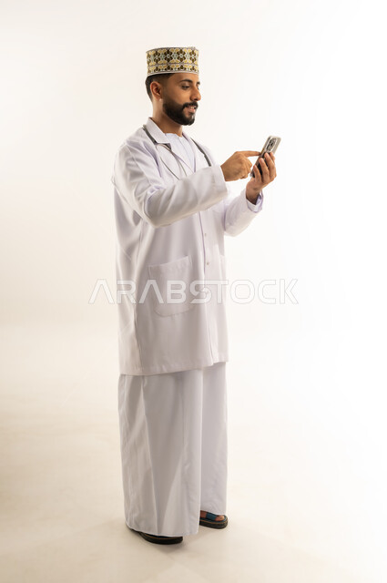 النقر بإصبع السبابة على شاشة الجوال، تقديم الاستشارة للمرضى عن بعد, بورتريه لطبيب عربي خليجي عماني يرتدي الدشداشة والكمة والزي الخاص بالمهنة يستخدم الهاتف المحمول، استخدام جهاز تقني حديث, العمل في القطاع الطبي في سلطنة عمان، الجسم بالطول الكامل، خلفية بيضاء