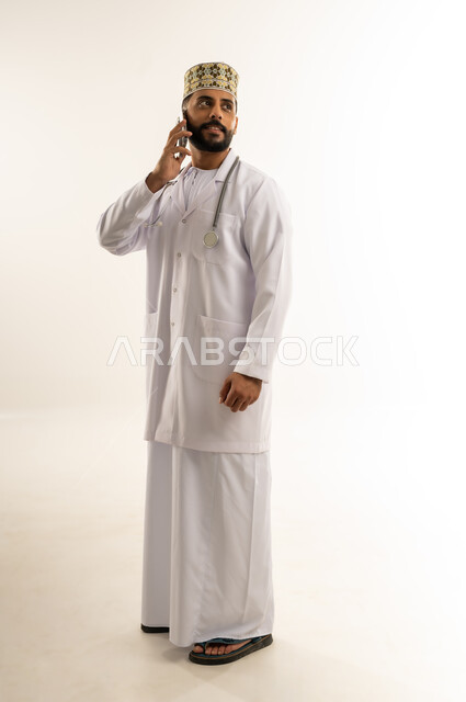 Providing remote consultation to patients via mobile phone, portrait of an Arab Gulf Omani doctor wearing a dishdasha, a kimono and a professional uniform making a phone call via a mobile phone looking at something above, using a modern technical device, full body, white background