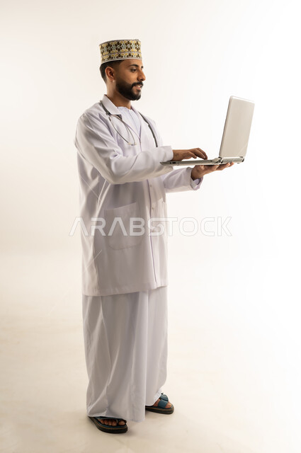 تقديم الاستشارة للمرضى عن بعد من خلال اللابتوب, بورتريه لطبيب عربي خليجي عماني يرتدي الدشداشة والكمة والزي الخاص بالمهنة يعمل على الحاسوب المحمول بإيماءات التركيز، العمل في القطاع الطبي في سلطنة عمان، استخدام جهاز تقني حديث، الجسم بالطول الكامل، خلفية بيضاء