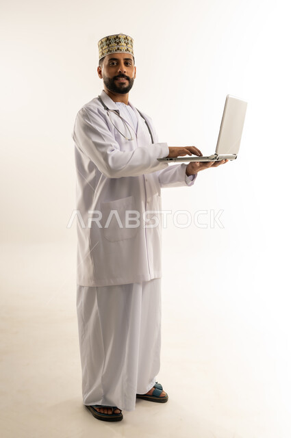 العمل في القطاع الطبي في سلطنة عمان، تقديم الاستشارة للمرضى عن بعد من خلال اللابتوب, بورتريه لطبيب عربي خليجي عماني يرتدي الدشداشة والكمة والزي الخاص بالمهنة يعمل على الحاسوب المحمول ينظر إلى الكاميرا، استخدام جهاز تقني حديث، الجسم بالطول الكامل، خلفية بيضاء