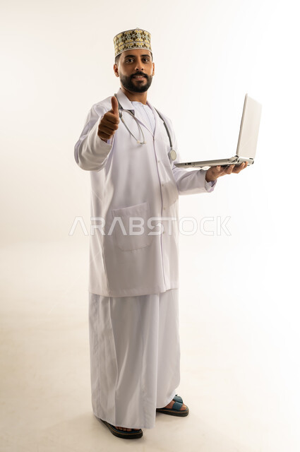 علامة الجودة والامتياز، استخدام جهاز تقني حديث، بورتريه لطبيب عربي خليجي عماني يرتدي الدشداشة والكمة والزي الخاص بالمهنة يمسك الحاسوب المحمول ينظر إلى الكاميرا يرفع إصبع الإبهام بإيماءات الإعجاب والقبول، العمل في القطاع الطبي، الجسم بالطول الكامل، خلفية بيضاء