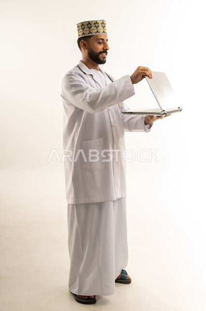 Using a modern technical device, providing consultation to patients remotely via laptop, portrait of an Arab Gulf Omani doctor wearing a dishdasha, a kimono and the professional uniform holding a laptop, working in the medical sector in the Sultanate of Oman, full body, white background