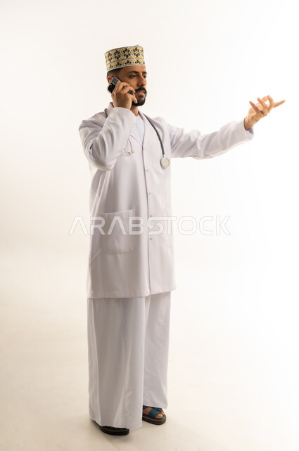 رفع اليد بإيماءات التساؤل والاستغراب، استخدام جهاز تقني حديث, تقديم الاستشارة للمرضى عن بعد من خلال الجوال, بورتريه لطبيب عربي خليجي عماني يرتدي الدشداشة والكمة والزي الخاص بالمهنة يقوم بإجراء مكالمة هاتفية عبر الهاتف المحمول، الجسم بالطول الكامل، خلفية بيضاء