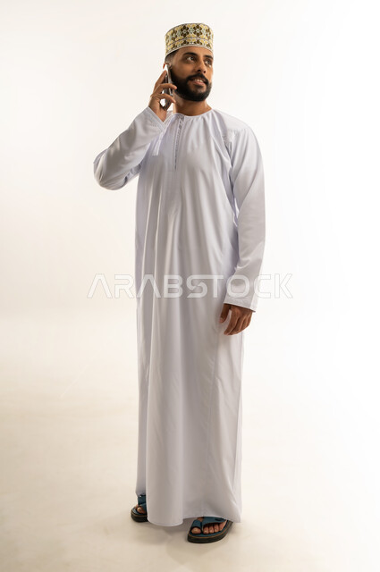 دمج التكنولوجيا المتطورة بالحياة اليومية، بورتريه لرجل عربي خليجي عماني يرتدي الدشداشة والكمة يقوم بإجراء مكالمة هاتفية عبر الهاتف المحمول, الدردشة والتواصل مع الأهل والأصدقاء وسماع أخبارهم، استخدام جهاز تقني حديث, الجسم بالطول الكامل، خلفية بيضاء