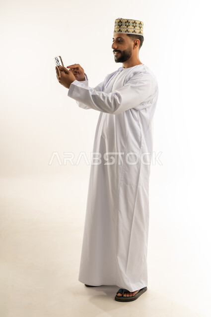 Using a modern technical device, integrating technology into daily life, portrait of an Arab Gulf Omani man wearing a dishdasha and a thobe using a mobile phone, tapping the index finger on the mobile screen, browsing social networking sites, chatting with family and friends, full body, white background