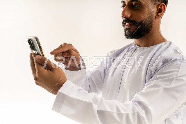 النقر بإصبع السبابة على شاشة الجوال، تصفح مواقع التواصل الاجتماعي، استخدام جهاز تقني حديث, دمج التكنولوجيا المتطورة بالحياة اليومية، بورتريه مقرب لرجل عربي خليجي عماني يرتدي الدشداشة يستخدم الهاتف المحمول, الدردشة مع الأهل والأصدقاء، خلفية بيضاء