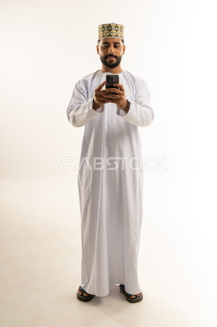 الدردشة مع الأهل والأصدقاء، تصفح مواقع التواصل الاجتماعي، استخدام جهاز تقني حديث, دمج التكنولوجيا المتطورة بالحياة اليومية، بورتريه لرجل عربي خليجي عماني يرتدي الدشداشة والكمة يستخدم الهاتف المحمول, الجسم بالطول الكامل, خلفية بيضاء