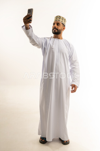 Taking a souvenir selfie, using a modern technical device, integrating advanced technology into daily life, portrait of an Arab Gulf Omani man wearing a dishdasha and a kummah holding a mobile phone in his hand taking a selfie with gestures of self-confidence, full body, white background