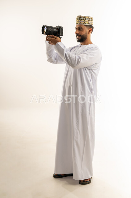 مصور محترف يلتقط صورة تذكارية، استخدام كاميرات رقمية عالية الدقة، بورتريه لرجل عربي خليجي عماني يرتدي الدشداشة والكمة يسمك بيده الكاميرا يقوم بالتقاط الصور الفوتوغرافية بإيماءات التركيز والتمعن، ممارسة هواية التصوير، الجسم بالطول الكامل، خلفية بيضاء