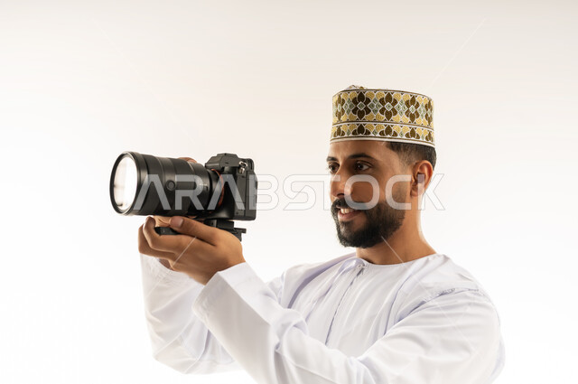 Professional photographer taking a souvenir photo, using high-resolution digital cameras, close-up portrait of an Arab Gulf Omani man wearing a dishdasha and a kummah holding a camera in his hand taking photographs with gestures of concentration and contemplation, practicing the hobby of photography, white background