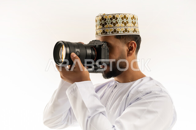 ممارسة هواية التصوير، مصور محترف يلتقط صورة تذكارية، استخدام كاميرات رقمية عالية الدقة، بورتريه مقرب لرجل عربي خليجي عماني يرتدي الدشداشة والكمة يسمك بيده الكاميرا يقوم بالتقاط الصور الفوتوغرافية بإيماءات التركيز والتمعن، خلفية بيضاء
