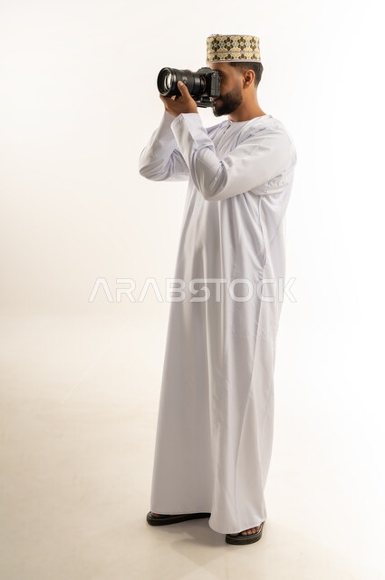 استخدام كاميرات رقمية عالية الدقة، بورتريه لرجل عربي خليجي عماني يرتدي الدشداشة والكمة يسمك بيده الكاميرا يقوم بالتقاط الصور الفوتوغرافية بإيماءات التركيز والتمعن، مصور محترف يلتقط صورة تذكارية، ممارسة هواية التصوير، الجسم بالطول الكامل، خلفية بيضاء