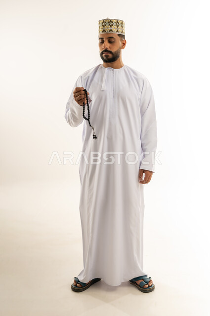 قراءة الأذكار والأدعية، التسبيح والتهليل وذكر الله تعالى، بورتريه لرجل عربي خليجي عماني يرتدي الدشداشة والكمة يمسك بيده السبحة، الاستغفار والصلاة على النبي الكريم، مفهوم العبادة والتقرب لرب العالمين، الجسم بالطول الكامل، خلفية بيضاء