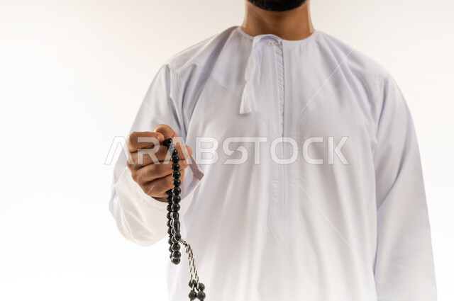 الاستغفار والصلاة على النبي الكريم، مفهوم العبادة والتقرب لرب العالمين، قراءة الأذكار والأدعية، التسبيح والتهليل وذكر الله تعالى، بورتريه مقرب لرجل عربي خليجي عماني يرتدي الدشداشة والكمة يمسك بيده السبحة، خلفية بيضاء